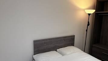 A neatly made bed with white linens, a bedside table with drawers, a floor lamp, and a wooden headboard.