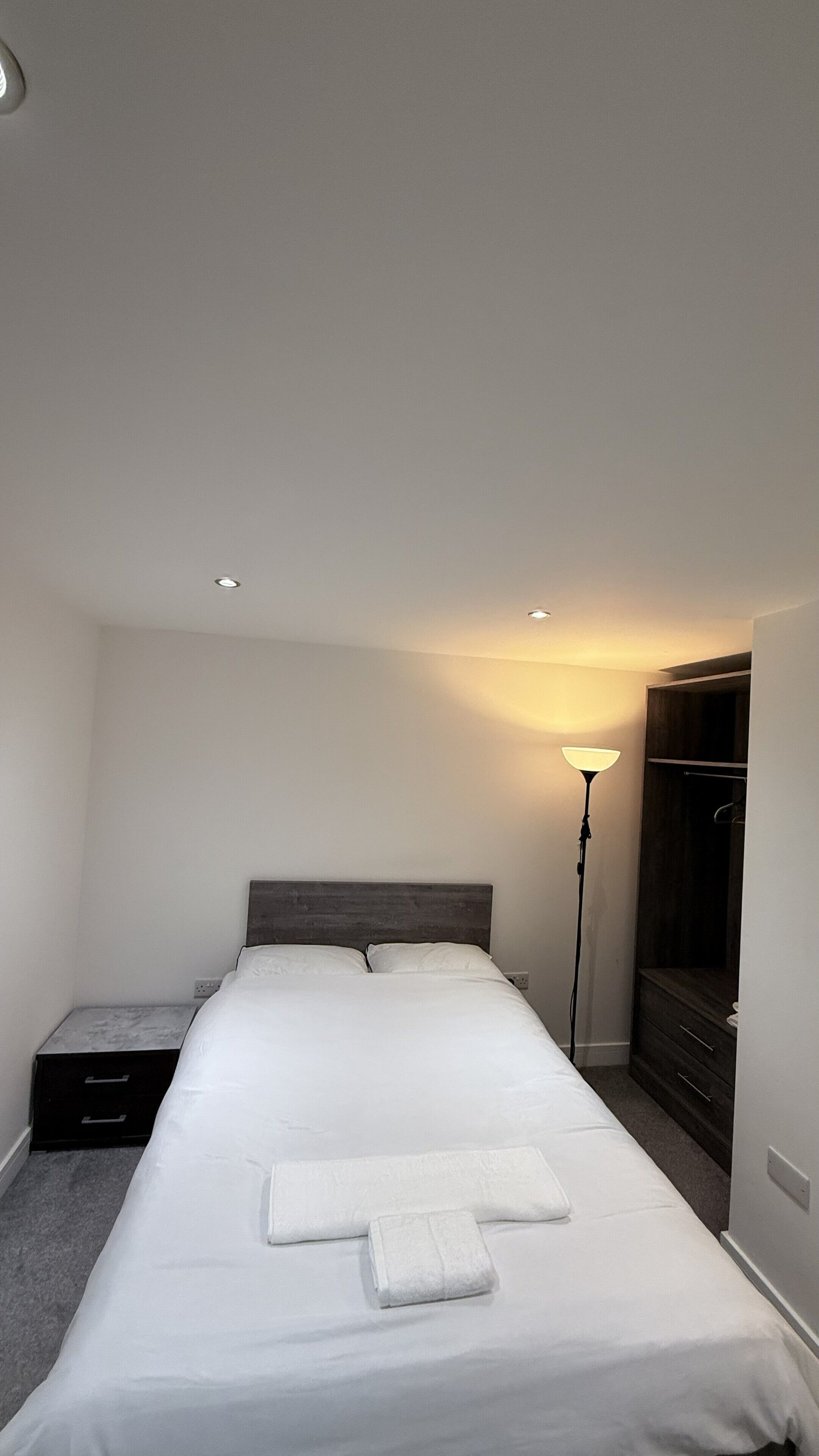 A neatly made bed with white linens and a folded towel, a bedside table, a lamp, and a wardrobe in a minimalistic room.