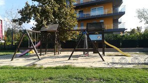 A playground with swings and slides, surrounded by a sandy area and a building in the background. - Studio with pool, hot tub and sauna near the free beach (Siófok)