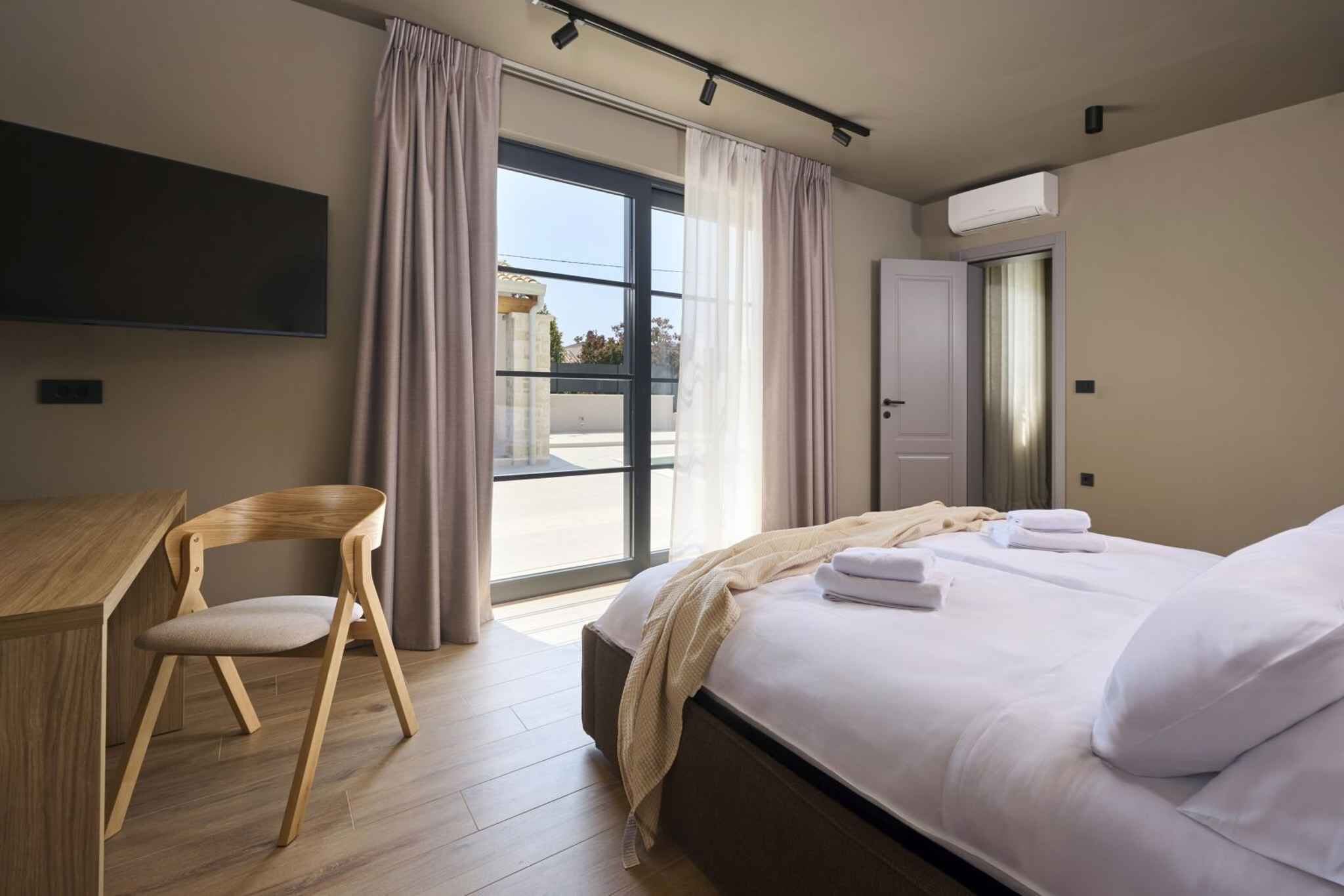 A modern hotel room with a bed, a desk, and a chair. There is a large window with curtains, an air conditioning unit, and a door.