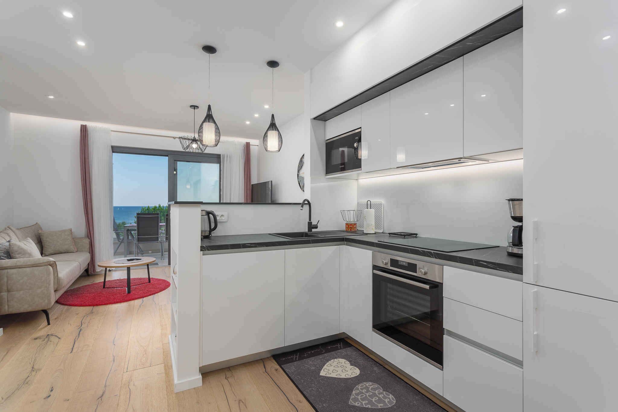 Una cocina moderna con armarios blancos, encimera negra y electrodomésticos de acero inoxidable. Hay un comedor con alfombra roja y vistas al exterior.