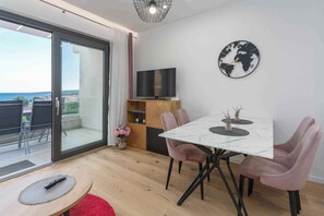 Un salon moderne doté d’une grande porte-fenêtre coulissante donnant sur un balcon, d’un téléviseur à écran plat fixé à un meuble en bois, d’une horloge mondiale ronde accrochée au mur et d’un coin repas avec une table en marbre et des chaises roses.