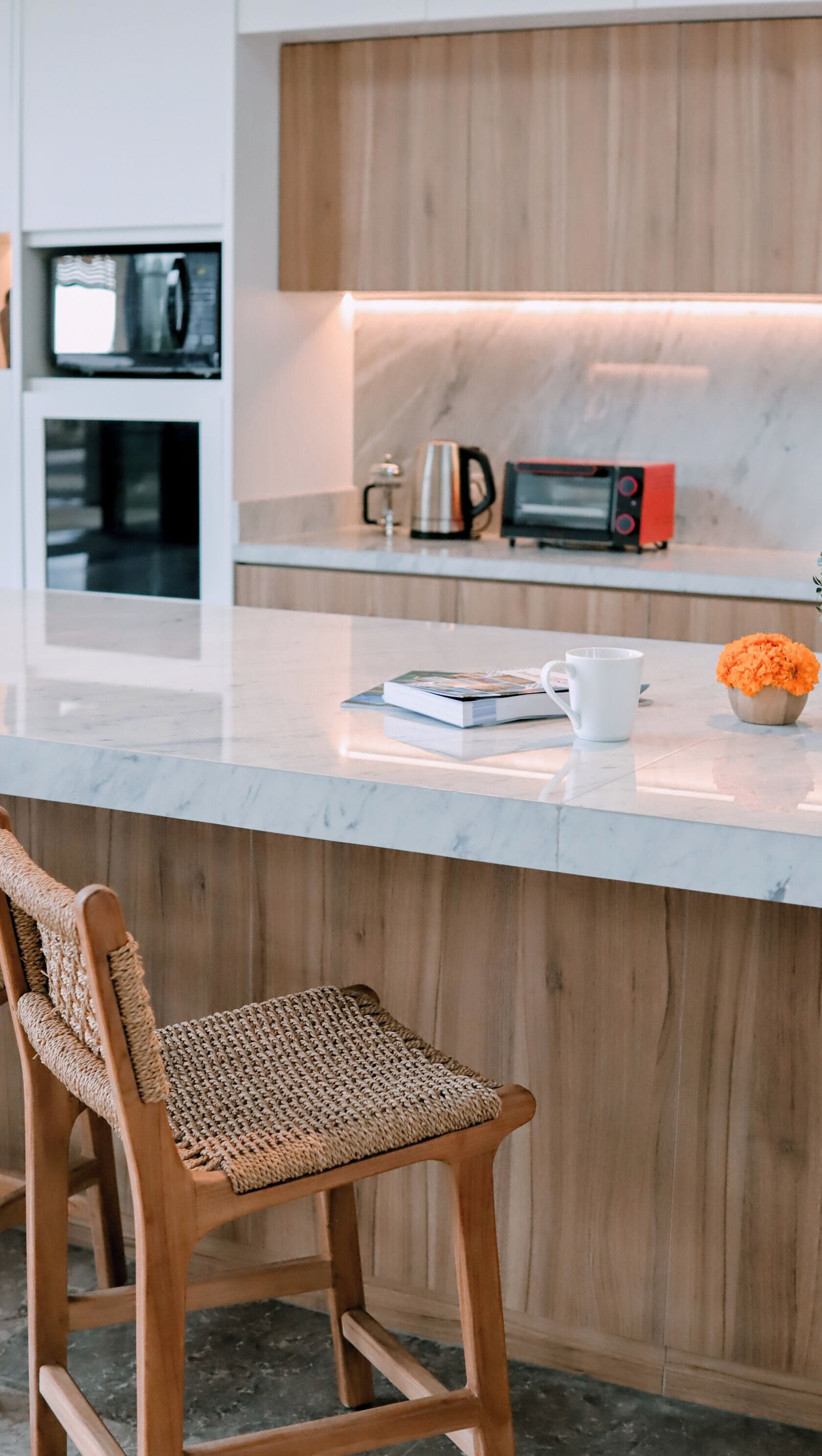 Una cocina con barra de madera, microondas, hervidor, y cafetera. Hay una silla tejida frente al mostrador.
