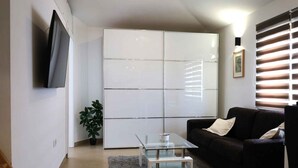 Una sala moderna con un televisor de pantalla plana montado en la pared, una mesa de centro de vidrio, un sofá de color oscuro y un mueble blanco grande.