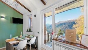 A dining area with a table set for two, a television mounted on the wall, and a view of mountains through a large window. - La Chicchetta Balcony & Mountain Views (Sauze d'Oulx)