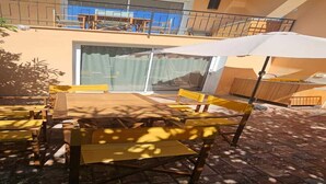 Un patio con bancos de madera y una sombrilla blanca, un balcón con barandilla, y un edificio con fachada amarilla.