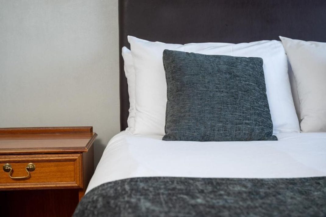A neatly made bed with white and gray bedding, a wooden nightstand with a drawer, and a dark headboard.