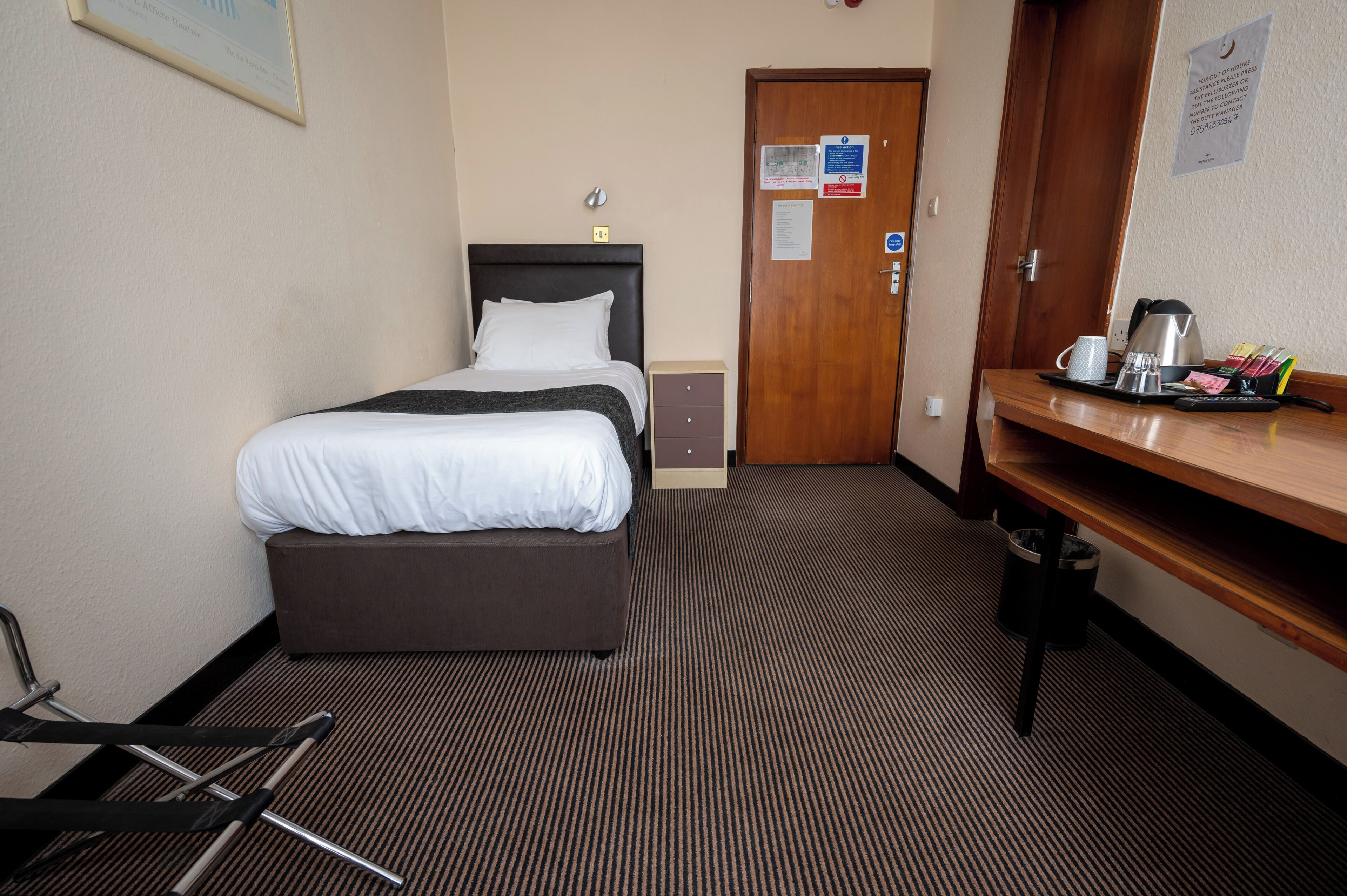 A hotel room with a bed, a desk with a kettle, and a trash bin.