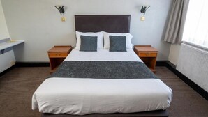 Une chambre d’hôtel avec un grand lit, deux tables de chevet, une petite table, une commode et une fenêtre avec des rideaux.
