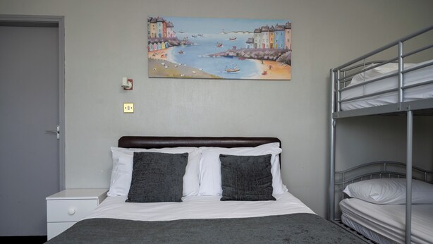 Une chambre avec un lit superposé, un lit simple avec une literie blanche, un plaid gris, un tableau représentant un paysage côtier et une table de chevet blanche avec un tiroir.