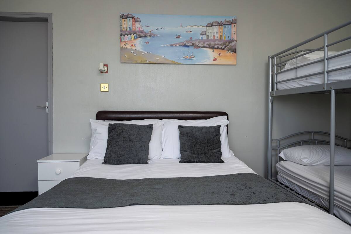 A bedroom with a bunk bed, a single bed with white bedding, a gray throw blanket, a painting of a coastal scene, and a white nightstand with a drawer.