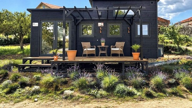 Een klein, donker houten huisje met een terras, twee stoelen en plantenpotten.