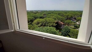 A room with a large window overlooking a dense forest and a distant view of a town. - Holiday flat with air conditioning and balcony with sea view (Lignano Sabbiadoro)