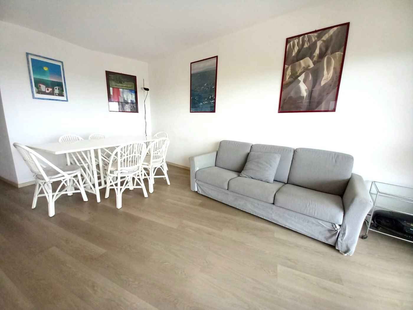 Ein Wohnzimmer mit grauem Sofa, einem weißen Esstisch mit Stühlen und drei gerahmten Bildern an der Wand.