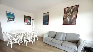 A living room with a grey sofa, a white dining table with chairs, and three framed pictures on the wall. - Holiday flat with air conditioning and balcony with sea view (Lignano Sabbiadoro)