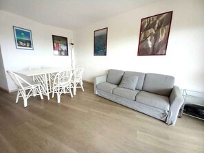 A living room with a grey sofa, a white dining table with chairs, and three framed pictures on the wall. - Holiday flat with air conditioning and balcony with sea view (Lignano Sabbiadoro)