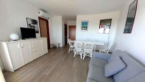 A living room with a grey sofa, a dining area with white chairs, a white cabinet with a TV, and a door leading to another room. - Holiday flat with air conditioning and balcony with sea view (Lignano Sabbiadoro)