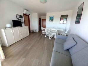 A living room with a grey sofa, a dining area with white chairs, a white cabinet with a TV, and a door leading to another room. - Holiday flat with air conditioning and balcony with sea view (Lignano Sabbiadoro)