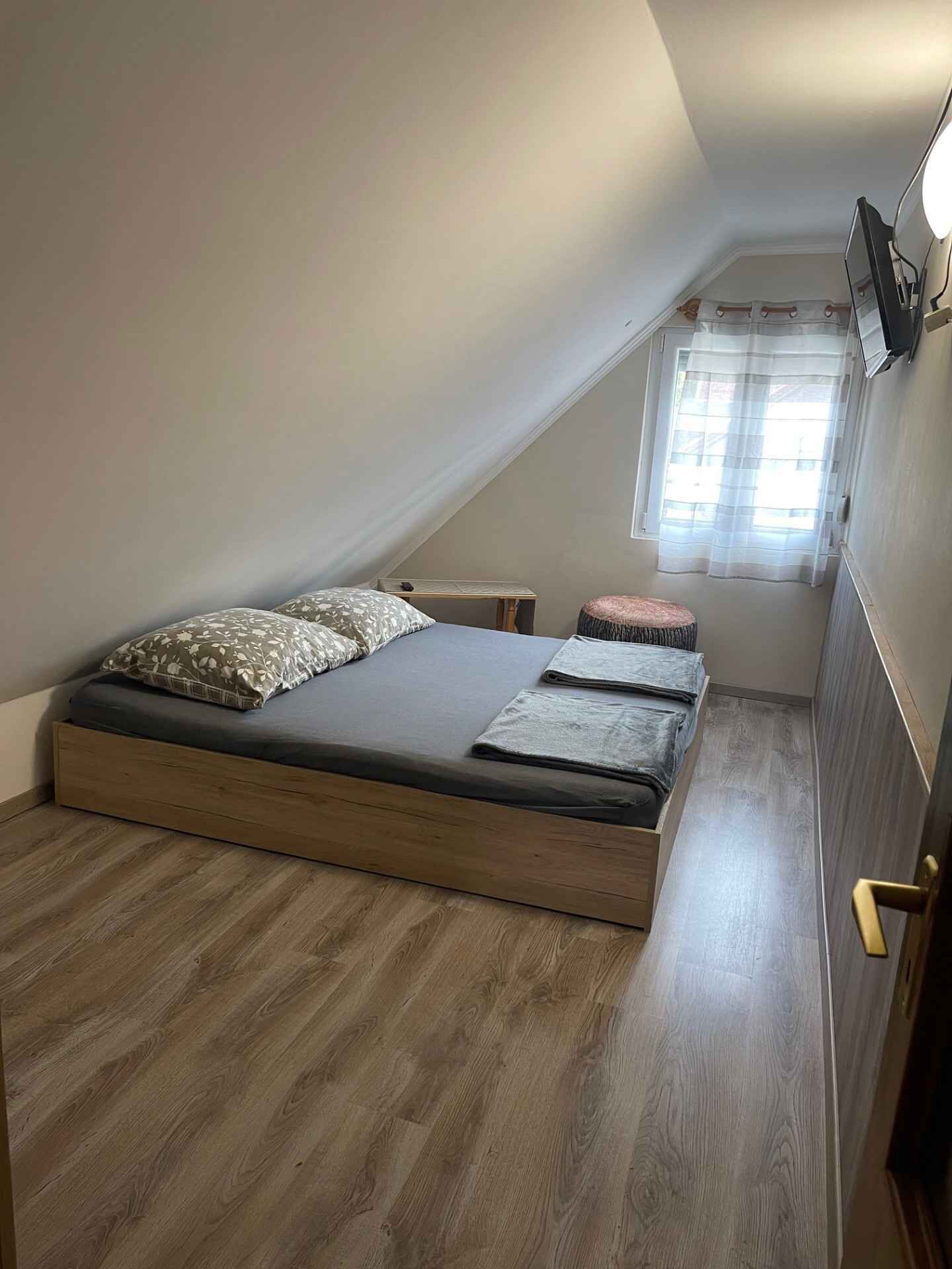 A small, sloped room with a single bed, a small table, and a window with sheer curtains.