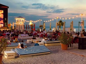 Eine Strandveranstaltung mit Liegestühlen und Sonnenschirmen, einer Bühne und Lichterketten.