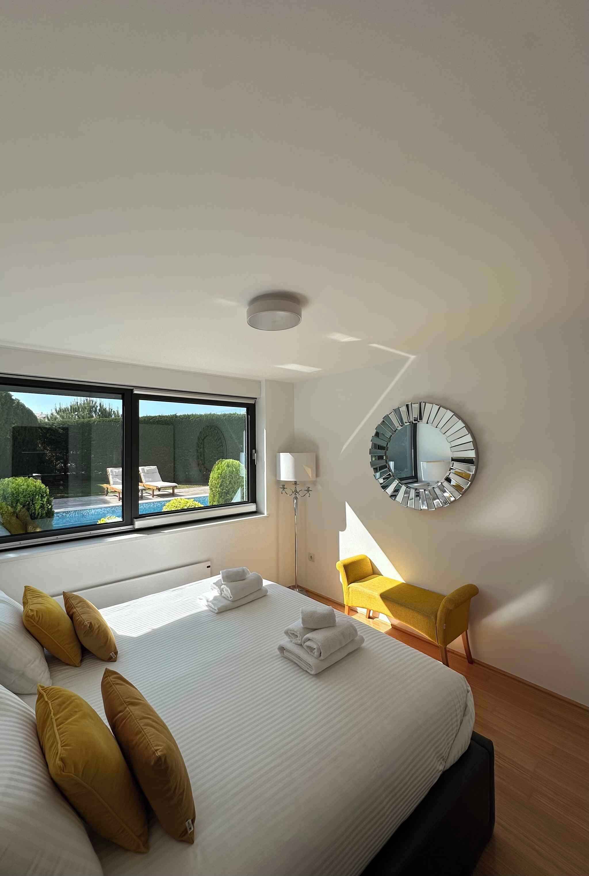 A hotel room with a large bed, a round mirror, a yellow chair, and a view of an outdoor swimming pool.