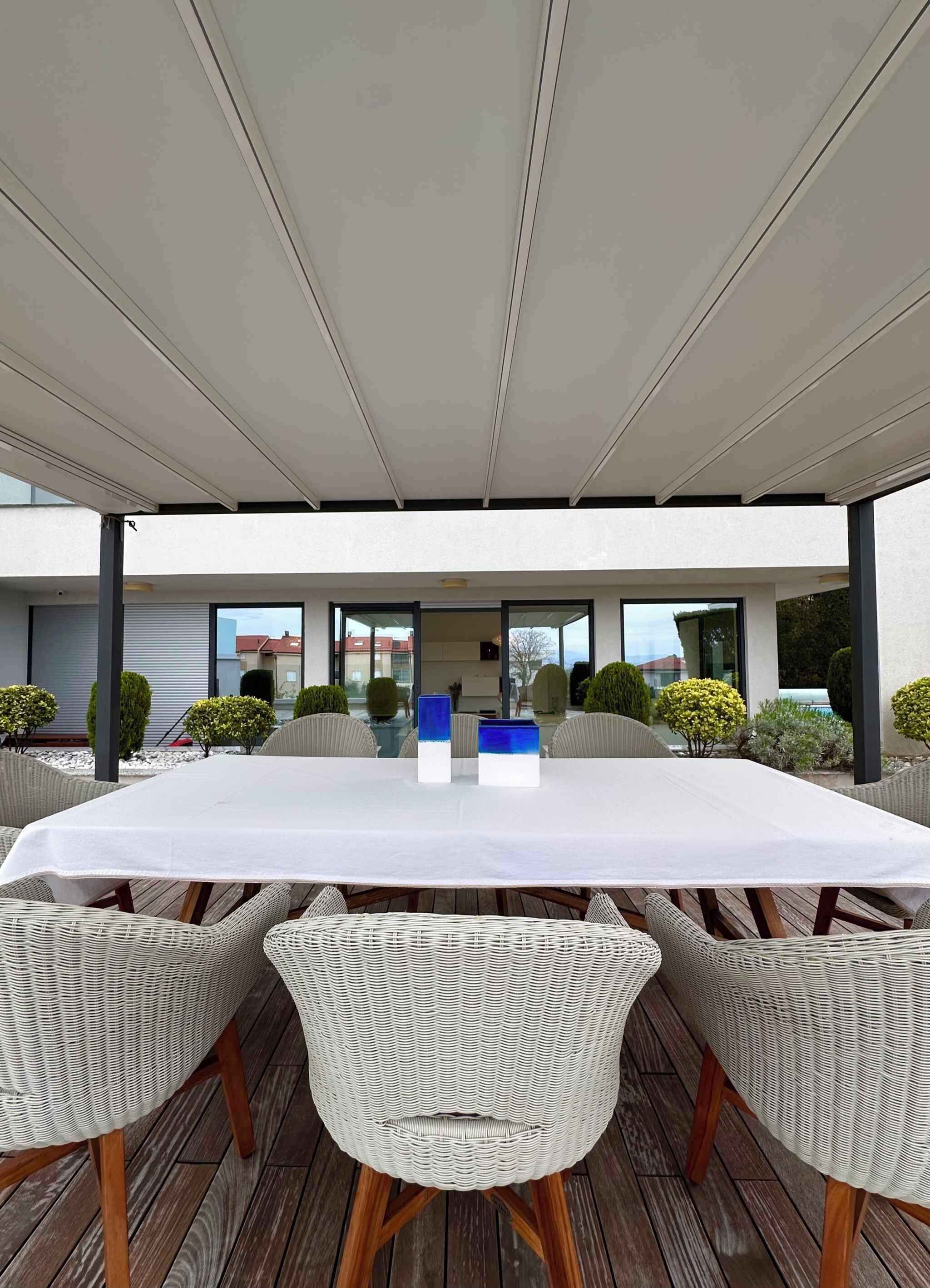 A covered outdoor dining area with a long table and wicker chairs, surrounded by greenery and a modern building in the background.