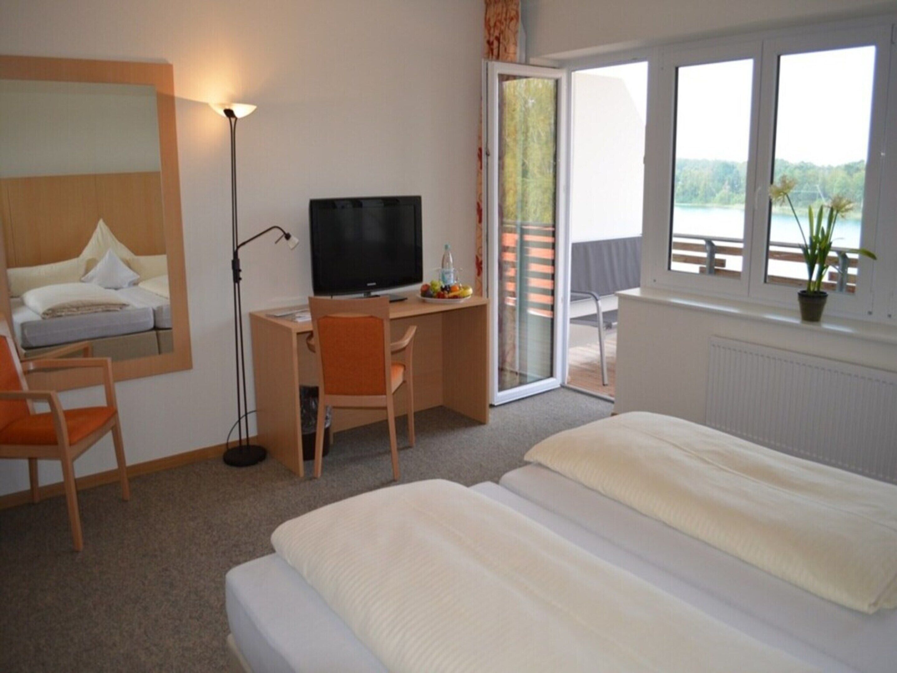 A hotel room with a bed, a desk with a computer, a chair, a TV, and a sliding glass door leading to a balcony with a view.