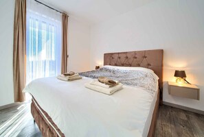 Een net opgemaakt bed met een gestoffeerd hoofdeinde, een bedlampje en een raam met gordijnen.