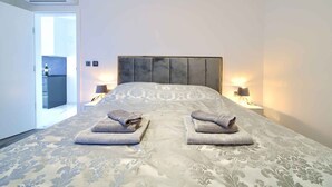 Ein Hotelzimmer mit einem großen Bett, zwei Nachttischlampen und Blick in die Küche durch eine geöffnete Tür.