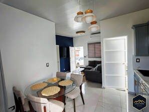 A modern kitchen with grey cabinets, a dining area with a glass table and cushioned chairs, and a living area with a dark sofa and a TV.