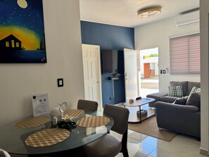 A modern living room with a grey sofa, a glass dining table with placemats, and a painting of a house under a starry sky.