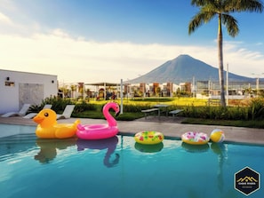 A swimming pool with inflatable toys, a mountain in the background, and a modern building with a white facade.