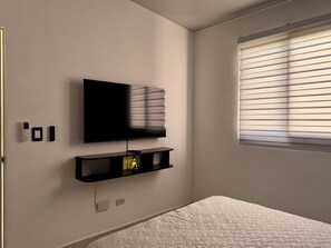 A bedroom with a bed, a mounted TV, a shelf with a lamp, and a window with blinds.