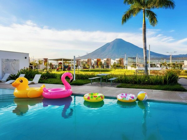 A swimming pool with inflatable toys, a palm tree, and a mountain in the background.