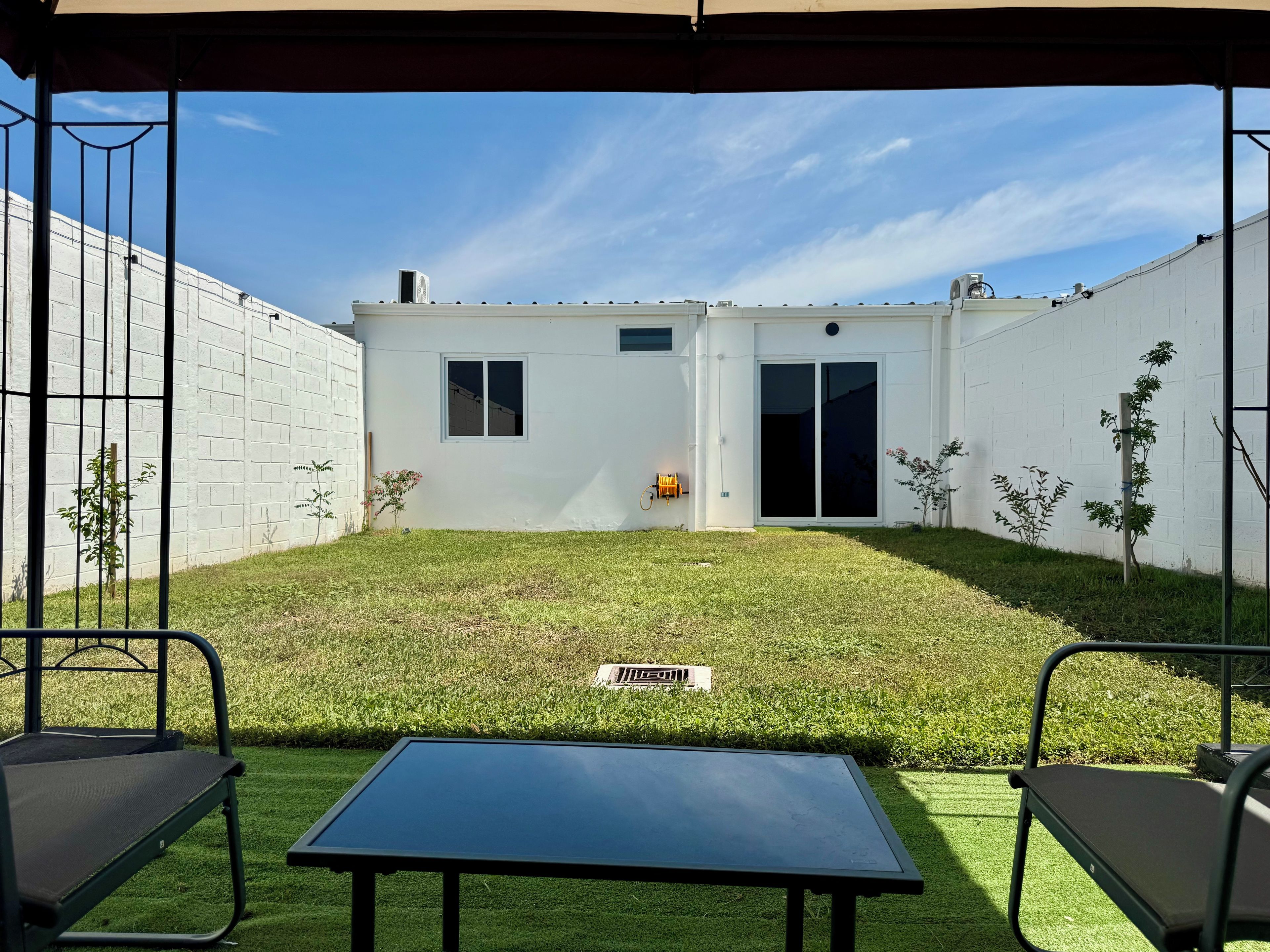 Un patio couvert avec une table et des chaises, un bâtiment blanc doté d’une porte-fenêtre coulissante et un petit jardin avec de la pelouse et des plantes.