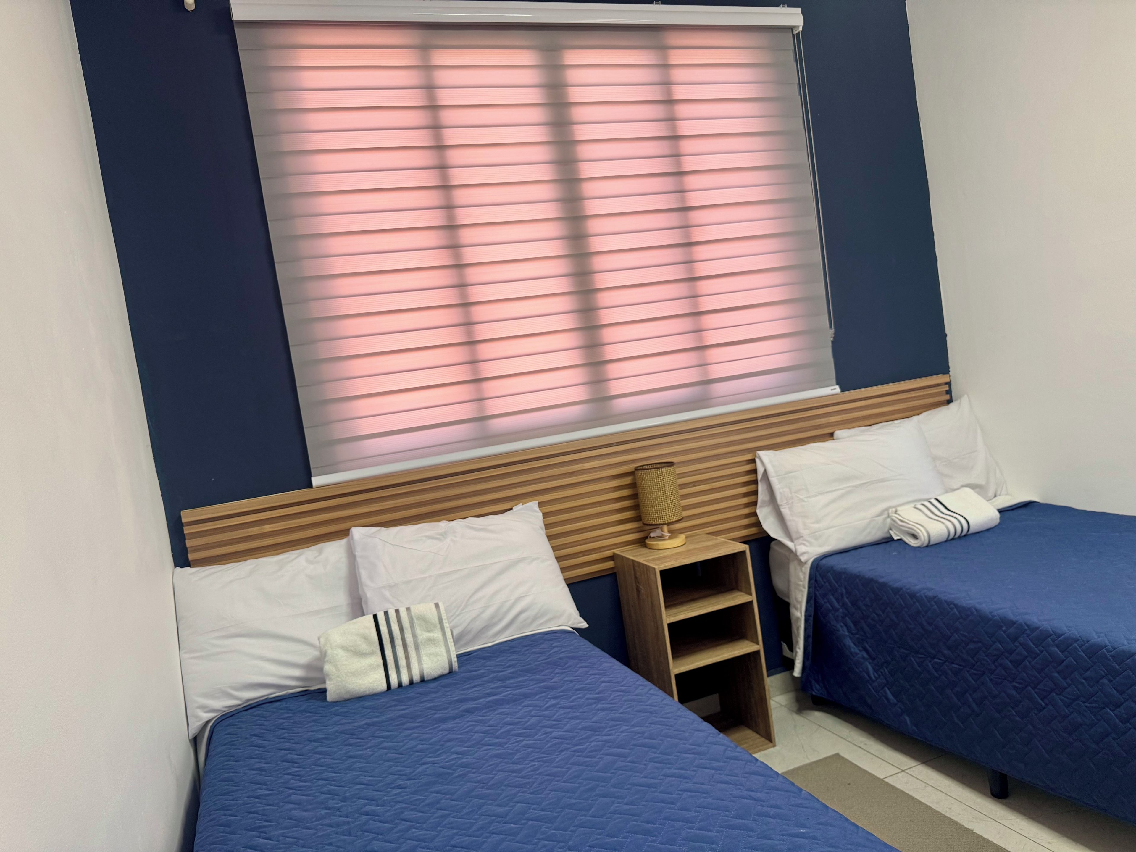 A room with two beds, each with a blue quilted bedspread, white pillows, and a wooden headboard. A small wooden nightstand with a lamp is between the beds.