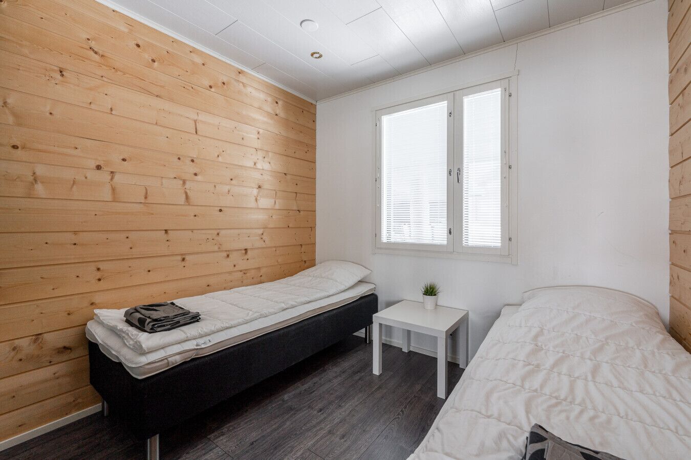 A small, single-room accommodation with two beds, a small table, and a window with blinds.