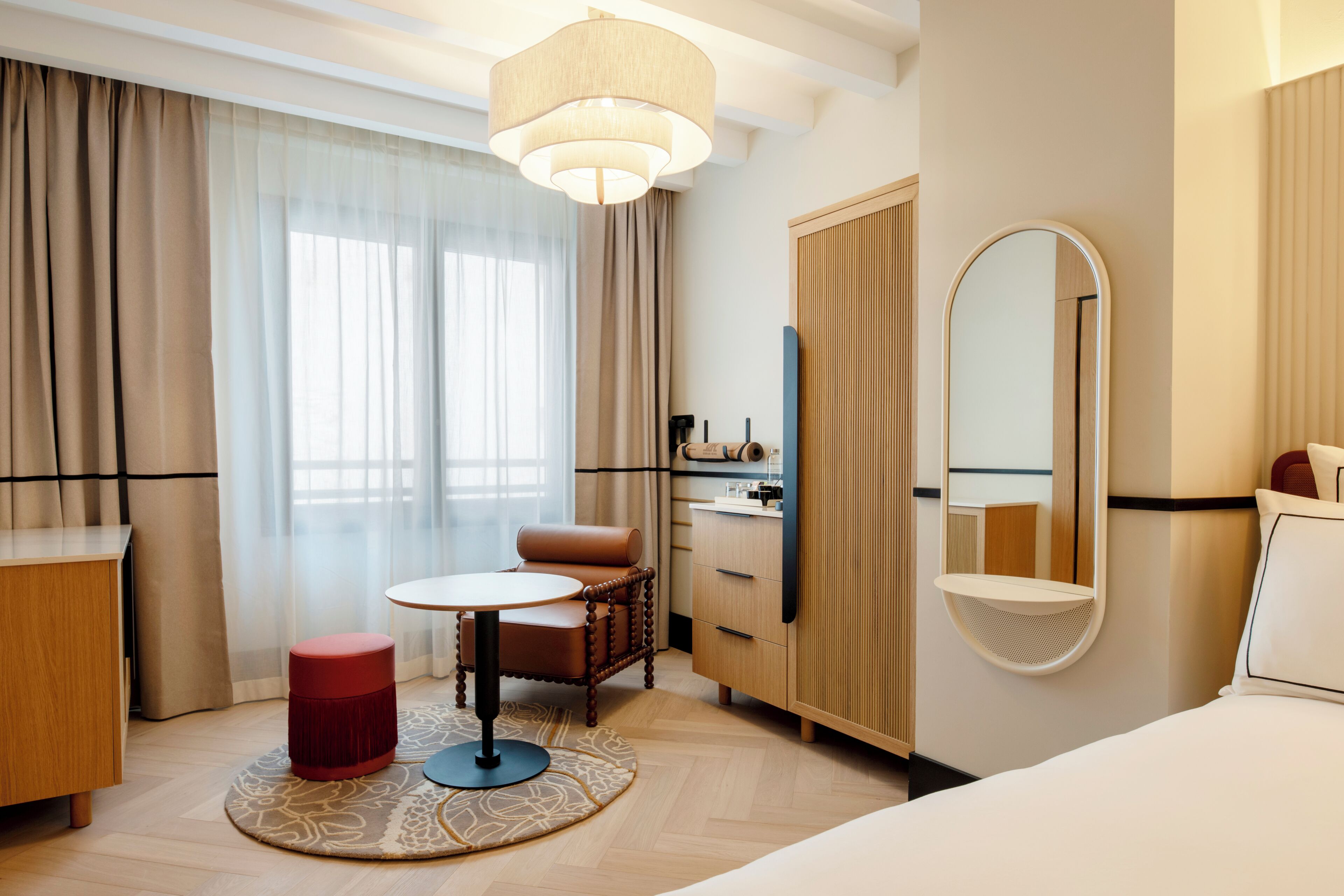 A hotel room with a bed, a round table, a chair, a wardrobe, a mirror, and a window with curtains.