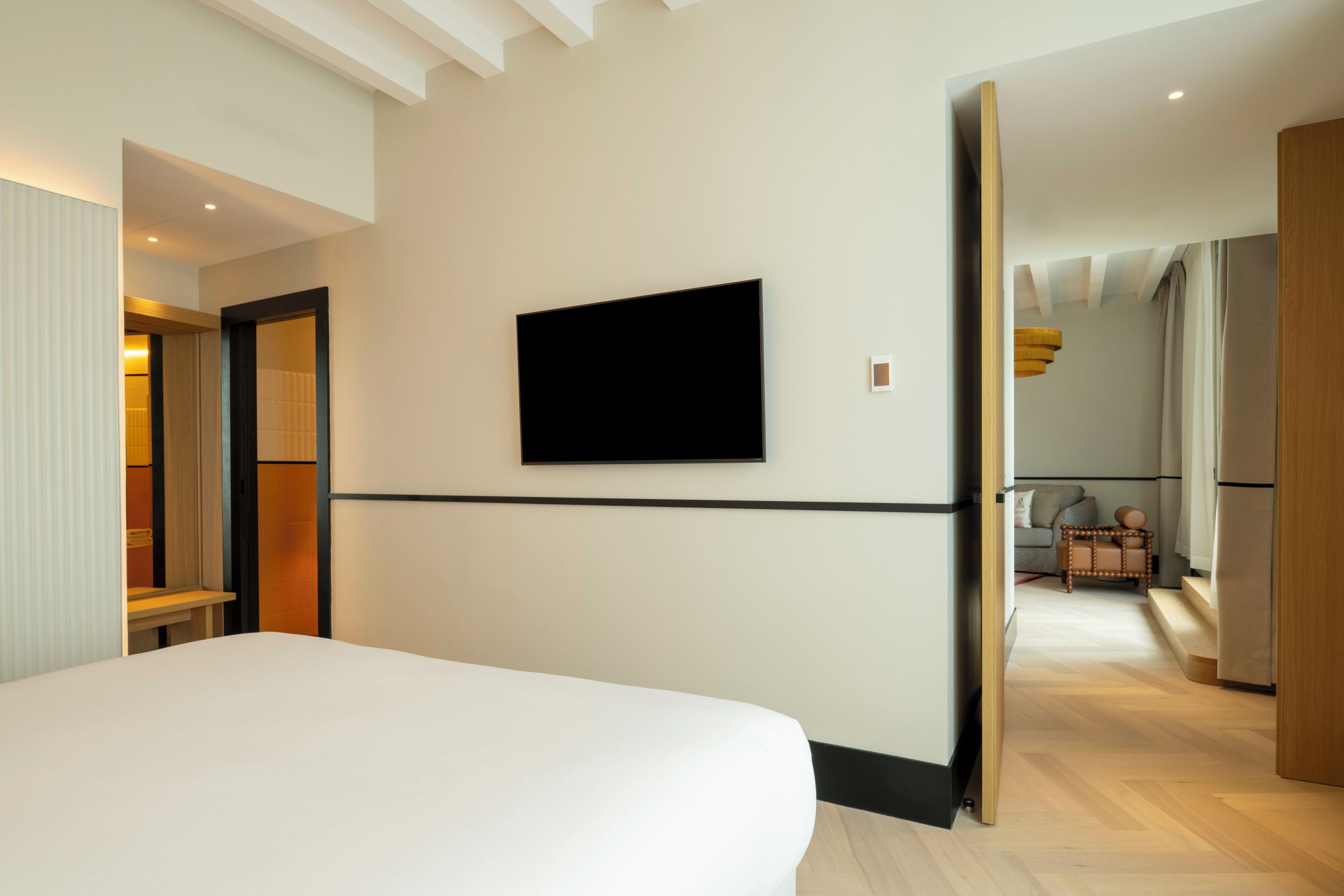 A modern hotel room with a large bed, a flat-screen TV mounted on the wall, and a wooden bench in the corner.