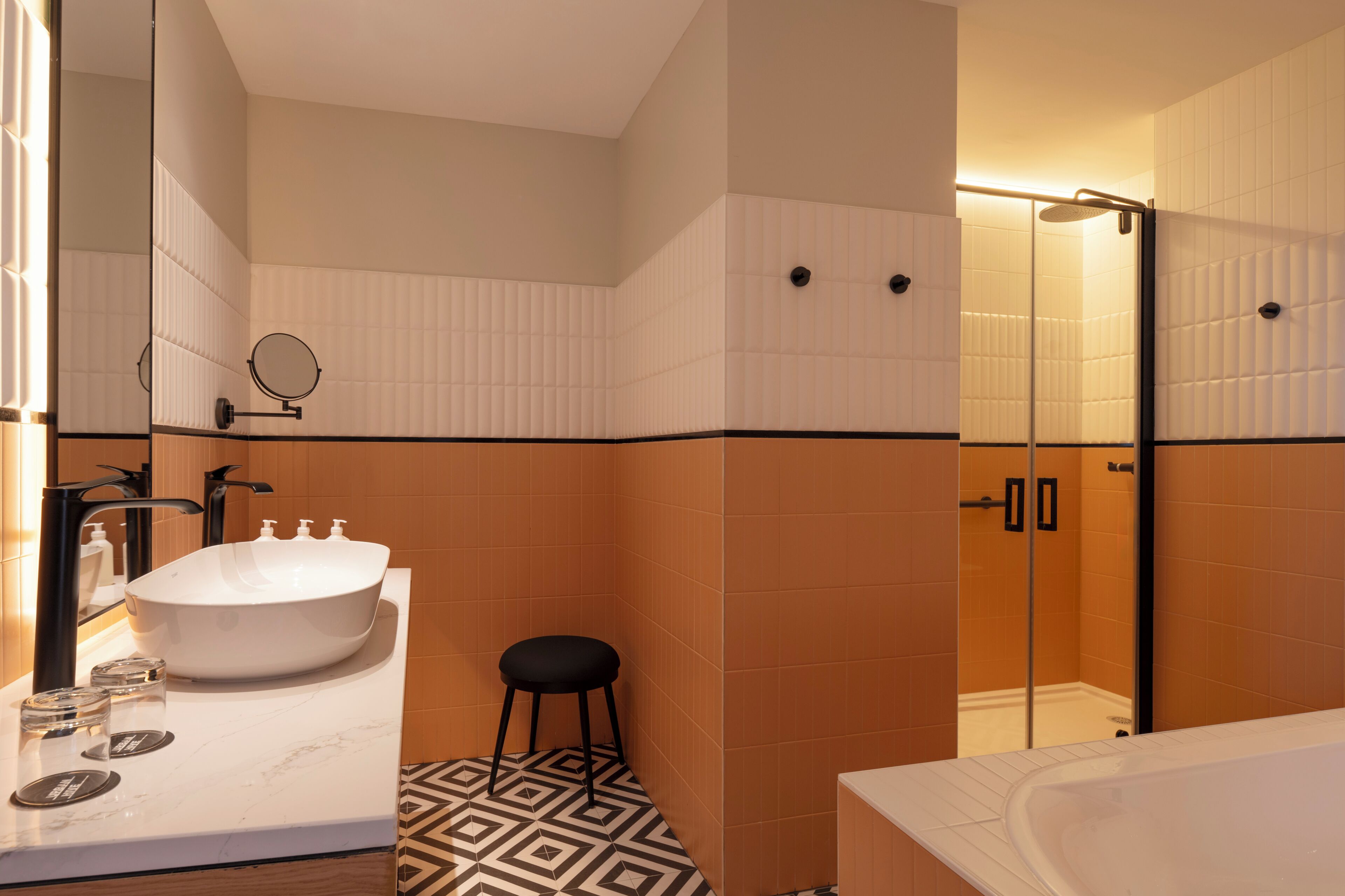 A bathroom with a large white sink, a round mirror, a black stool, a glass shower enclosure, and a patterned floor.