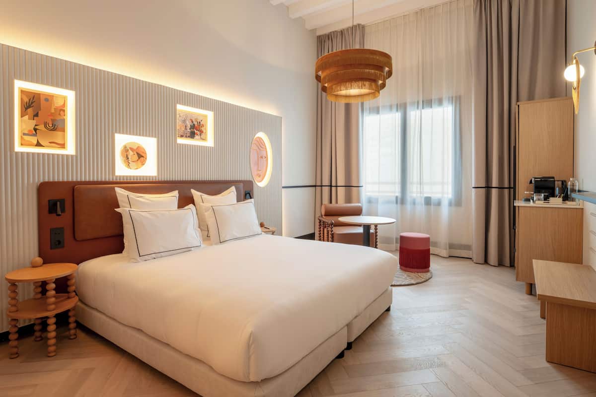 Ein modernes Hotelzimmer mit einem großen Bett, einem runden Tisch, einem kleinen Hocker, einem Schreibtisch und einem Stuhl.