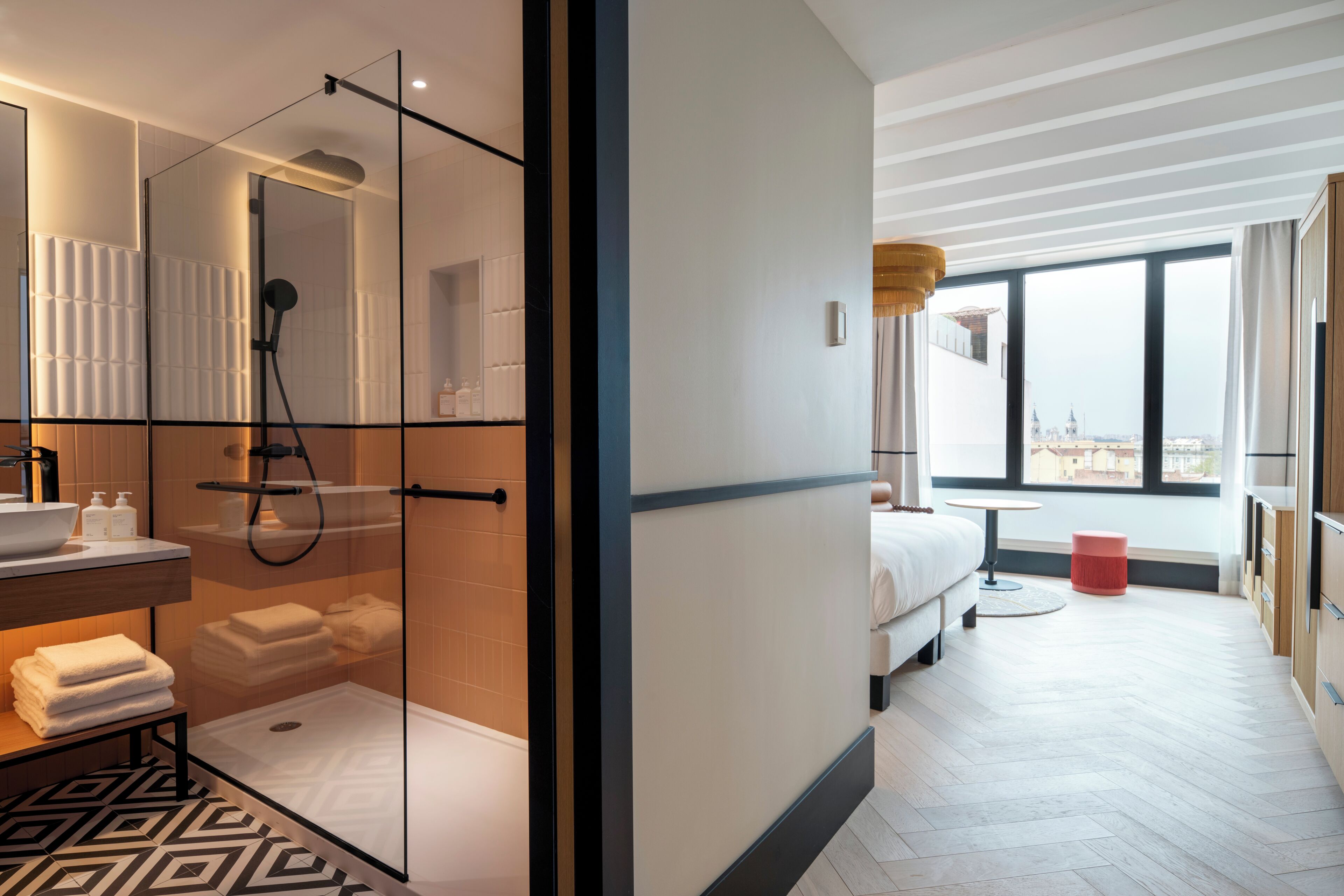Een moderne hotelkamer met een glazen douchecabine, een groot raam met uitzicht op de stad, een bed en een klein rond tafeltje.