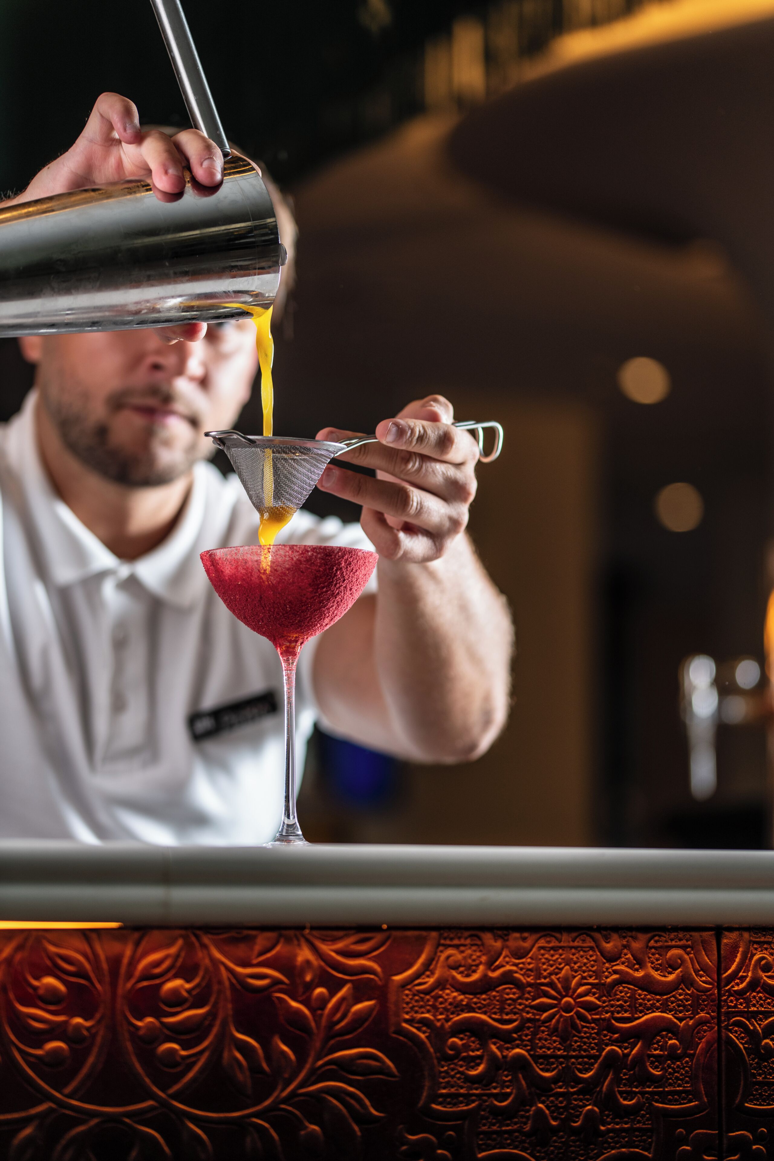 A bartender pouring a yellow liquid into a red cocktail glass.