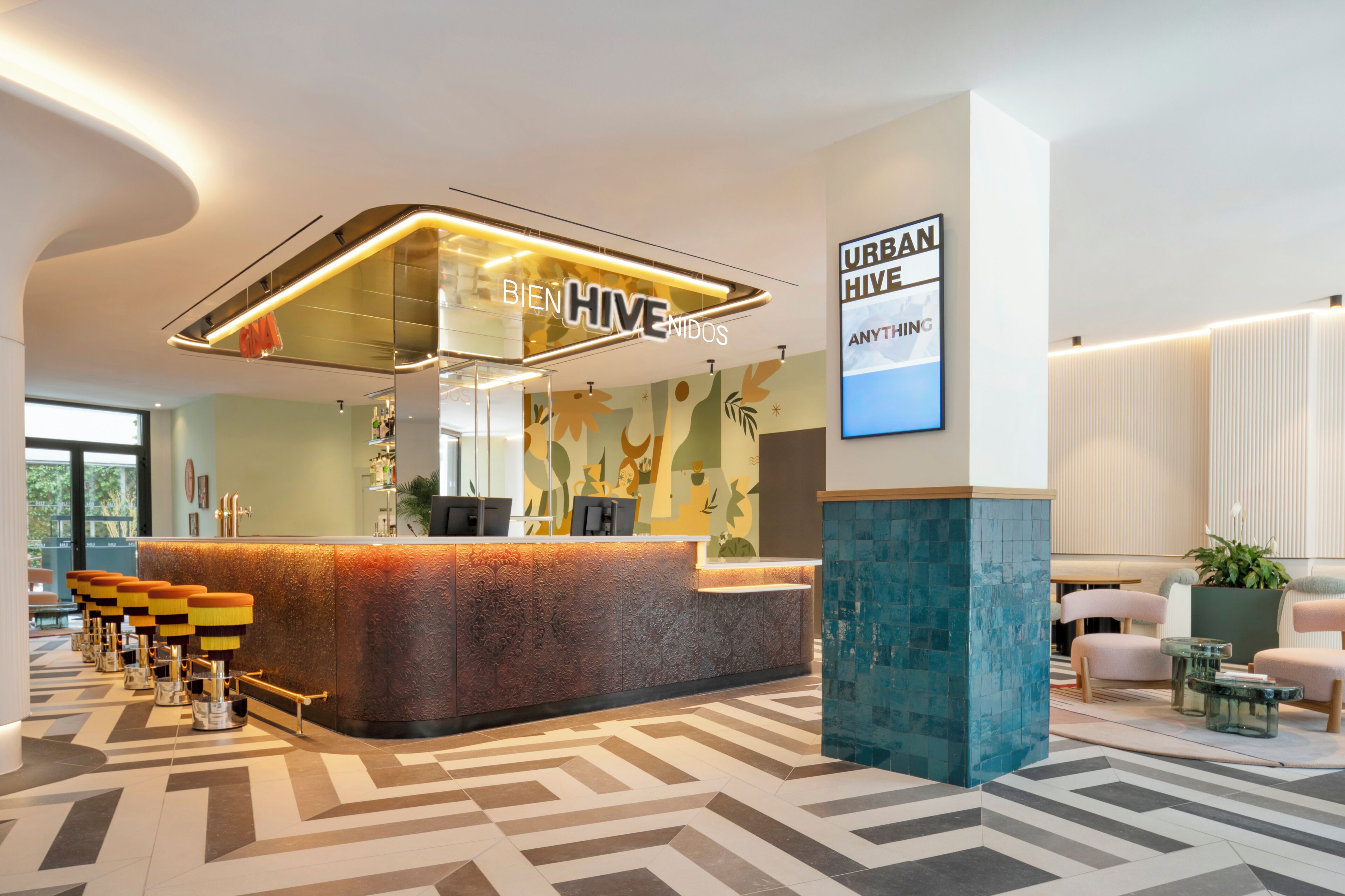 Ein moderner Empfangsbereich mit einer gebogenen Theke, einem Schild mit der Aufschrift 'BIEN HIVE NIDOS' und einer Wand mit dem Text 'URBAN HIVE ANYTHING'. Es gibt Sitzbereiche mit Tischen und Stühlen, sowie einen beflügelten Boden.