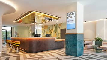 Ein moderner Empfangsbereich mit einer gebogenen Theke, einem Schild mit der Aufschrift 'BIEN HIVE NIDOS' und einer Wand mit dem Text 'URBAN HIVE ANYTHING'. Es gibt Sitzbereiche mit Tischen und Stühlen, sowie einen beflügelten Boden.