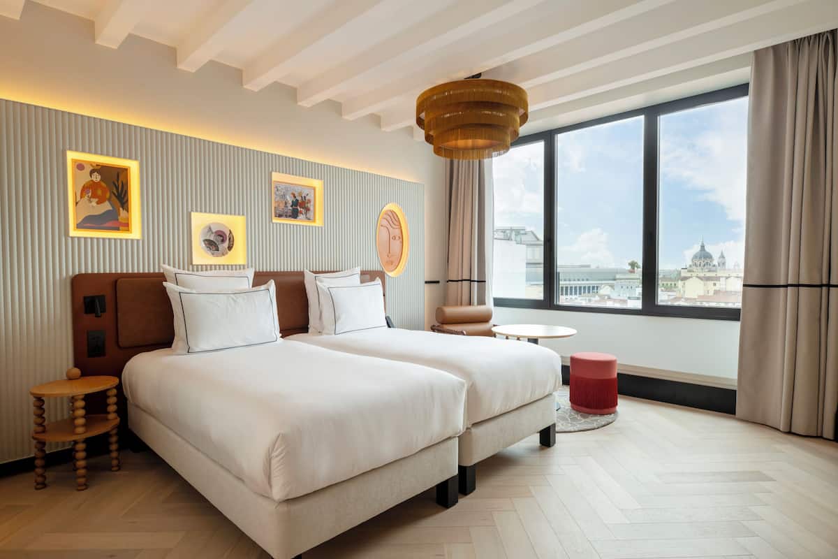 Ein Hotelzimmer mit einem großen Bett, einem kleinen runden Tisch, einem roten Hocker und Blick auf die Stadt durch große Fenster.