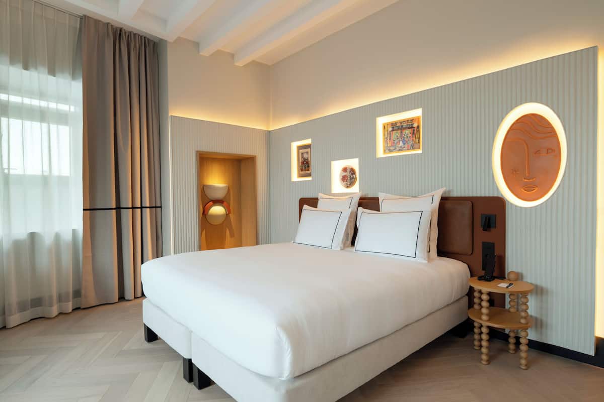 Ein Hotelzimmer mit einem großen Bett, einem Nachttisch, einer Wandlampe und gerahmten Kunstwerken.
