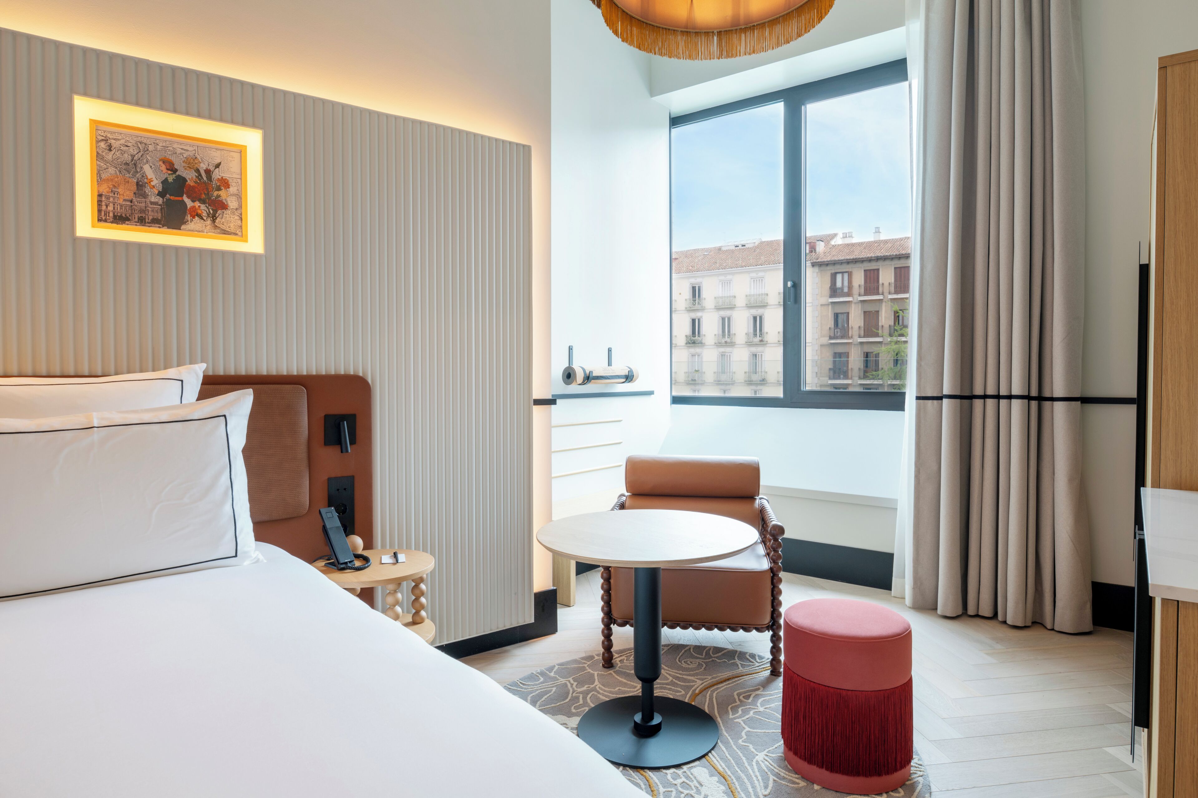 A hotel room with a bed, a round table, a chair, a small stool, a red ottoman, a window with curtains, a wall-mounted picture, and a view of buildings outside.