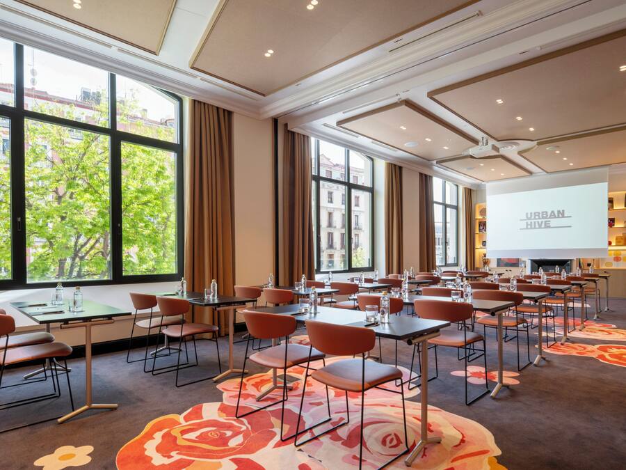 Une salle de conférence équipée de tables, de chaises, d’un écran de projection et de grandes fenêtres offrant une vue sur les bâtiments et les arbres.