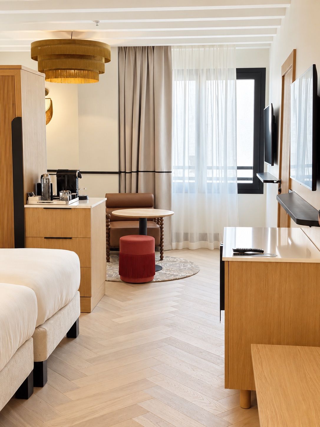 Una habitación de hotel moderna con cama, escritorio, silla, un puf rojo, televisión y un ventanal grande con cortinas.